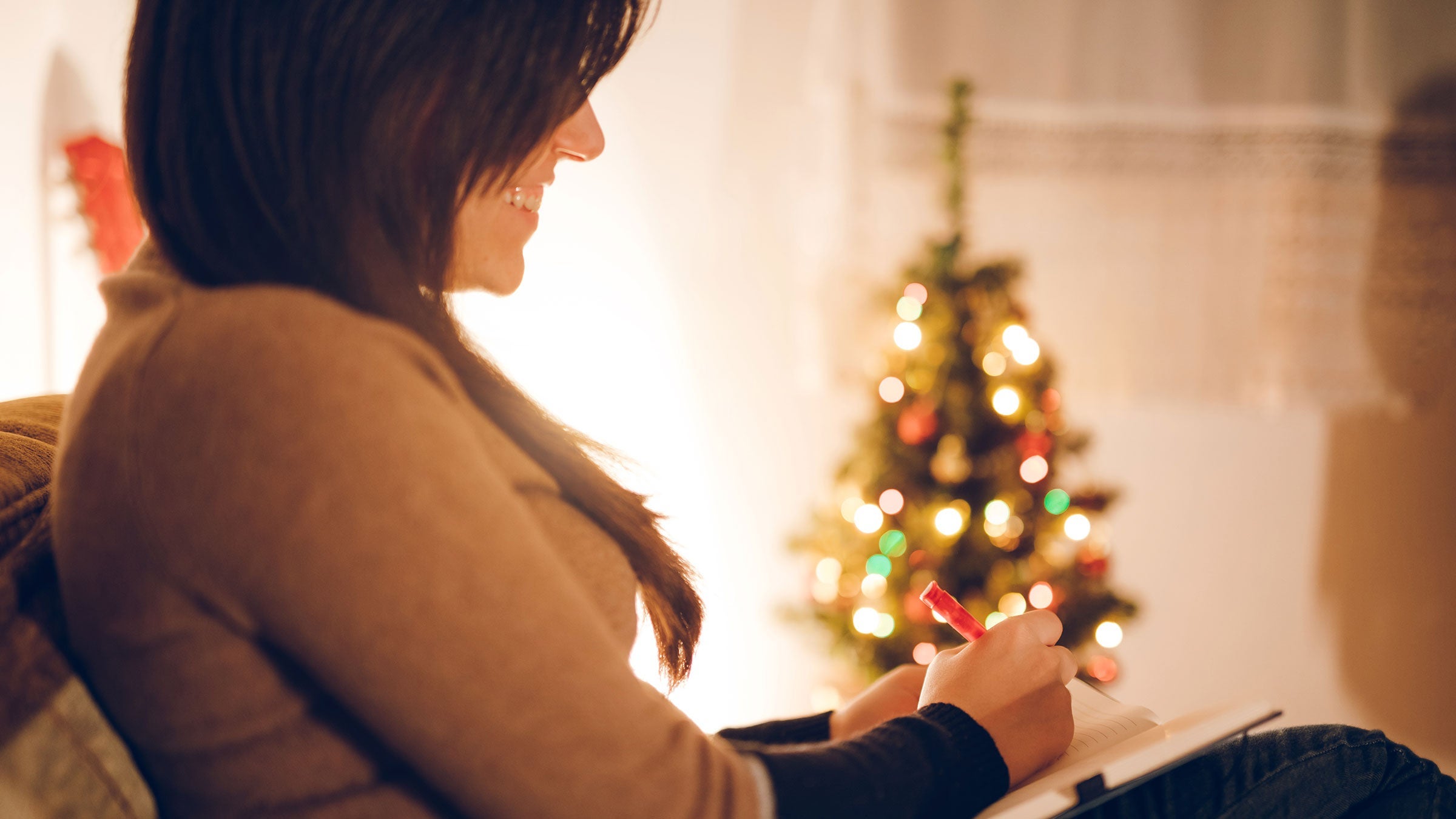 A woman writes in a journal while sitting near a holiday tree at home